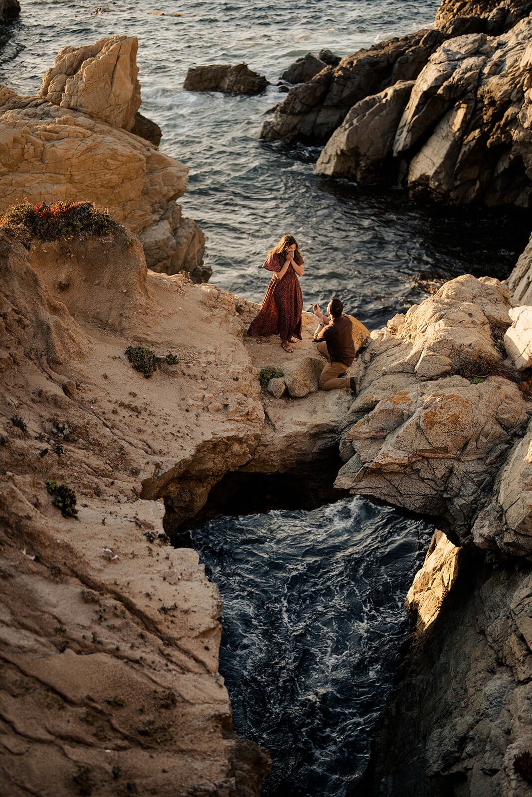Aerial view of a couple on rocky cliffs by the ocean, with the man kneeling to propose to the woman.