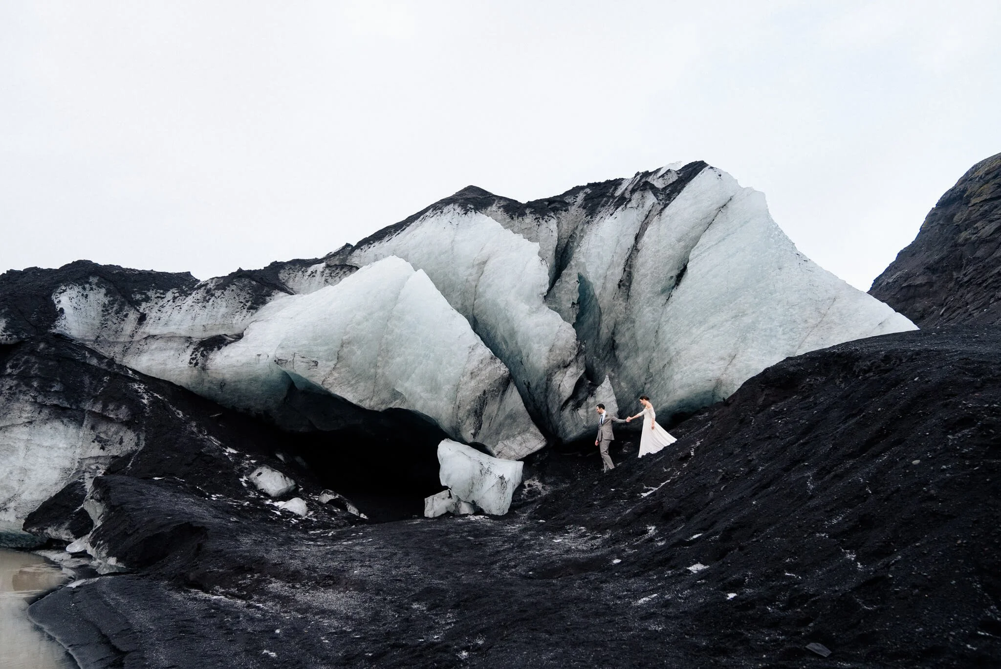 Bride and groom standing near a large glacier with dark rock surroundings.