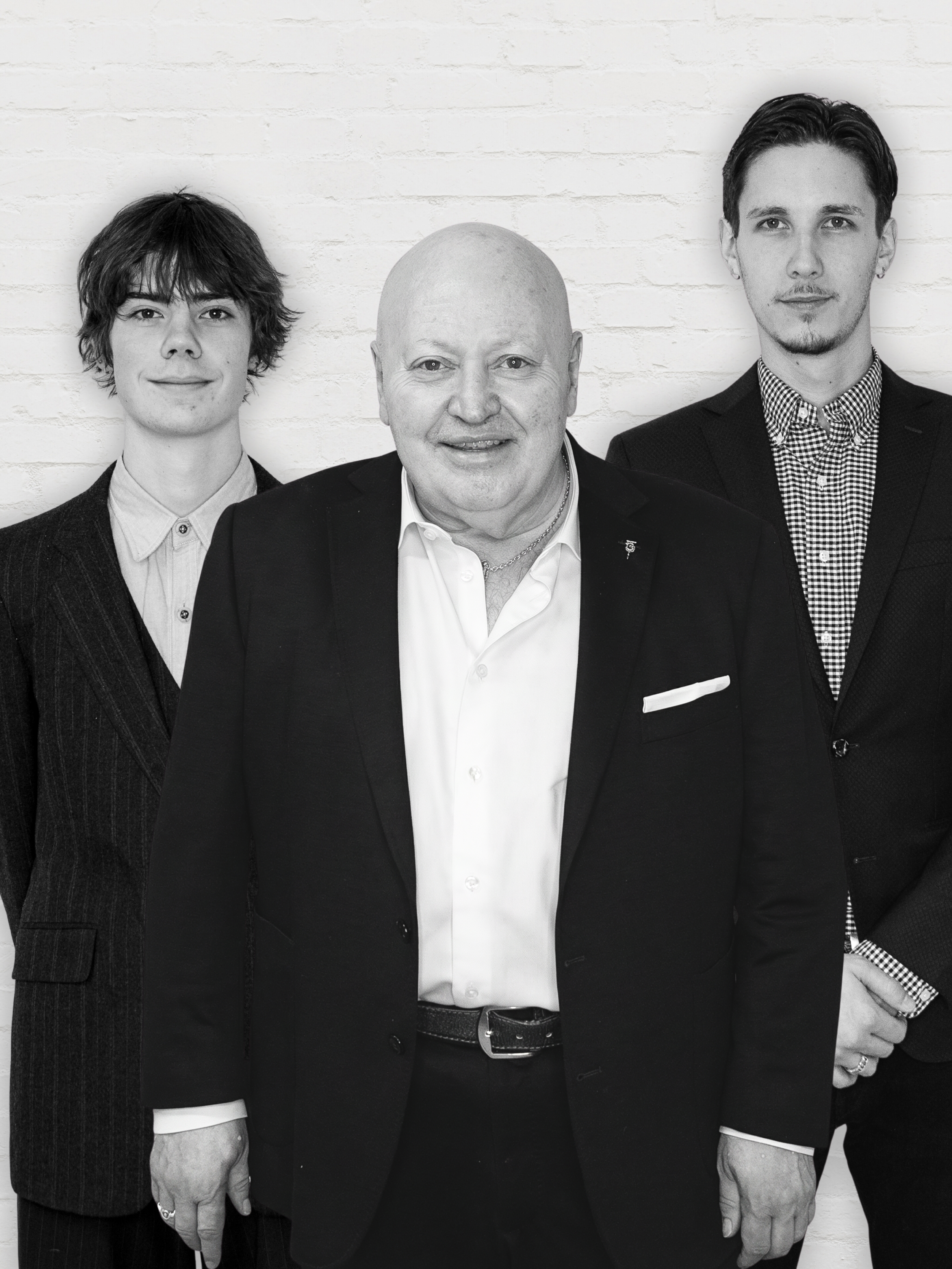 A person wearing a pinstripe suit, glasses, and a tie, standing with hands on hips in front of a white brick wall. Todd MartinPatrick 3