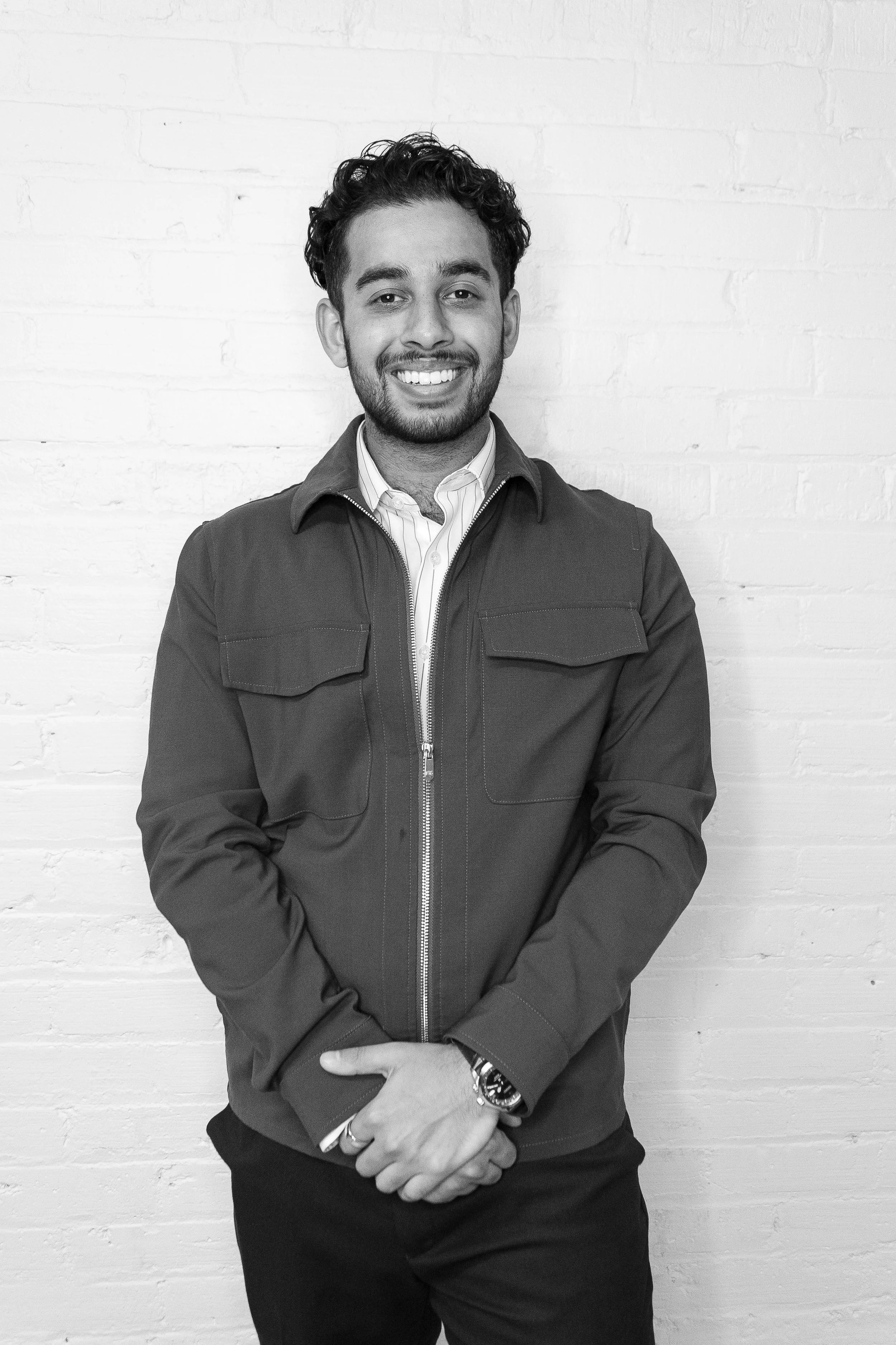 Man standing against a white brick wall, wearing a light blazer, white shirt, and dark pants, with hands in pockets, smiling. Ish MartinPatrick 3.