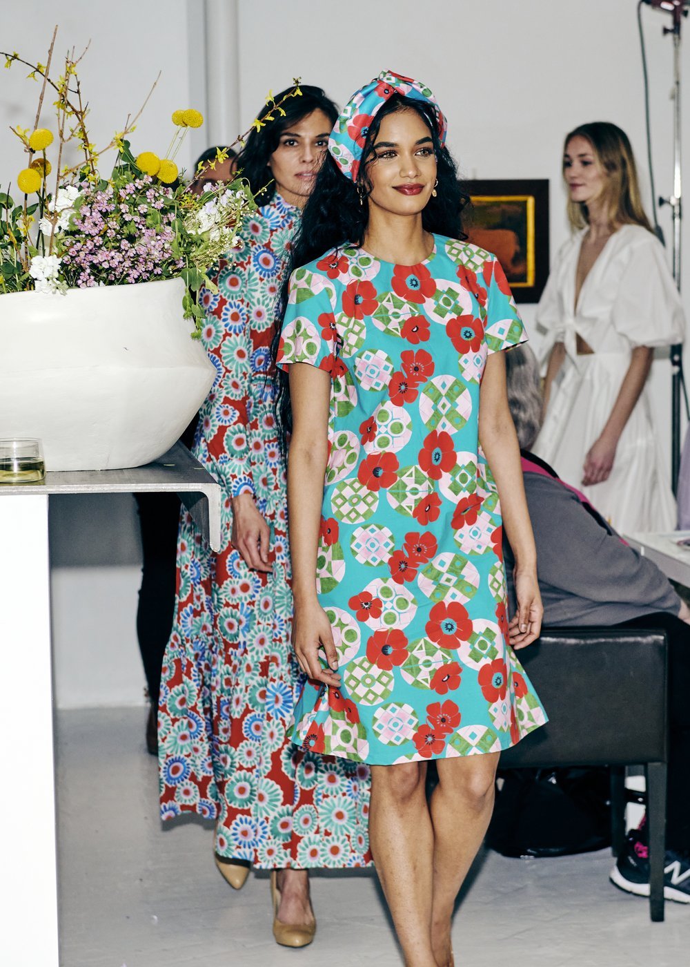 Two models wearing colorful floral dresses walk in a fashion show. One model also wears a matching headband. A large floral arrangement is visible in the foreground.