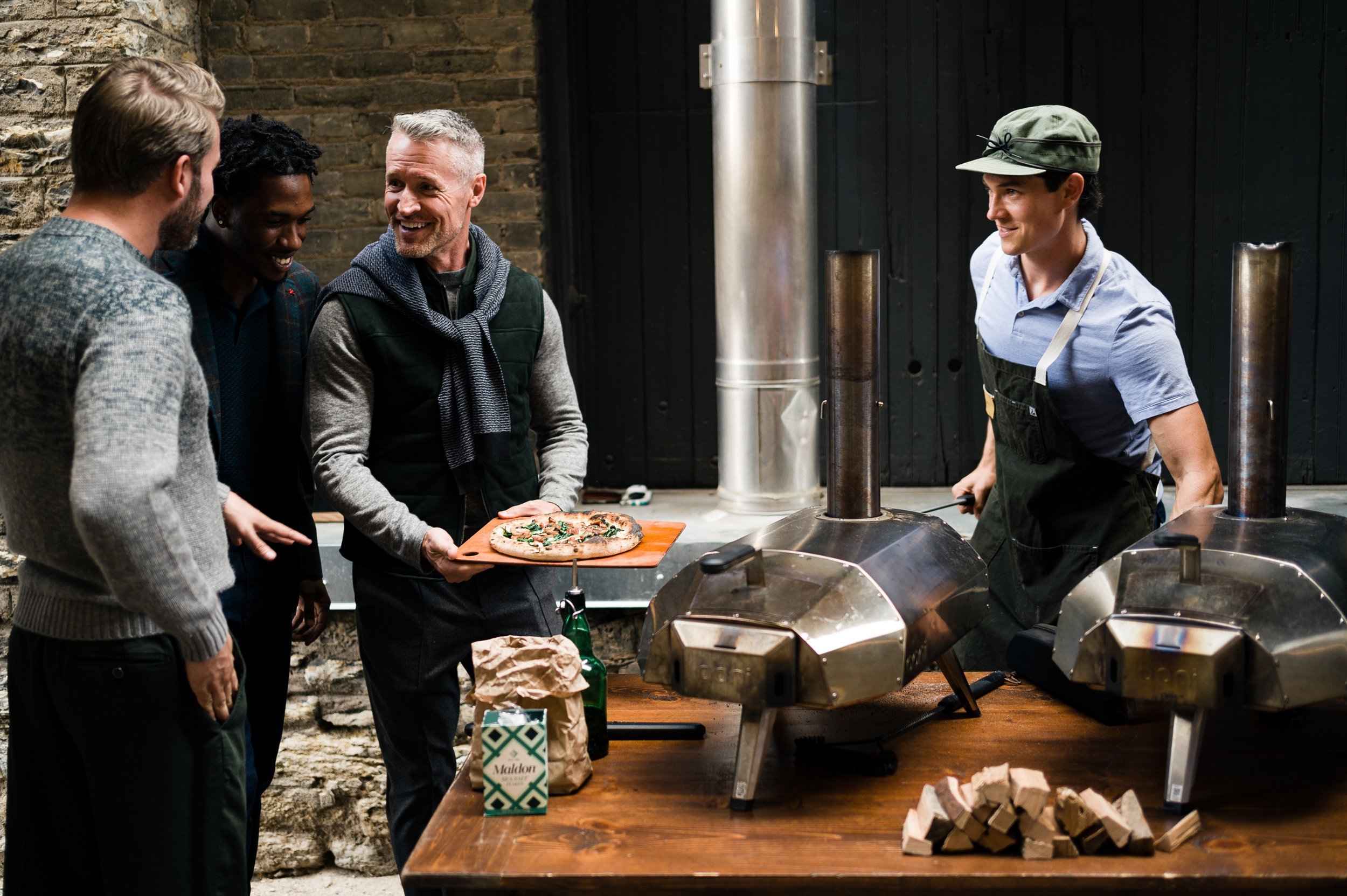People gathered around an outdoor pizza oven, one holding a pizza on a wooden board, with wood-fired ovens and cooking supplies visible on a table.