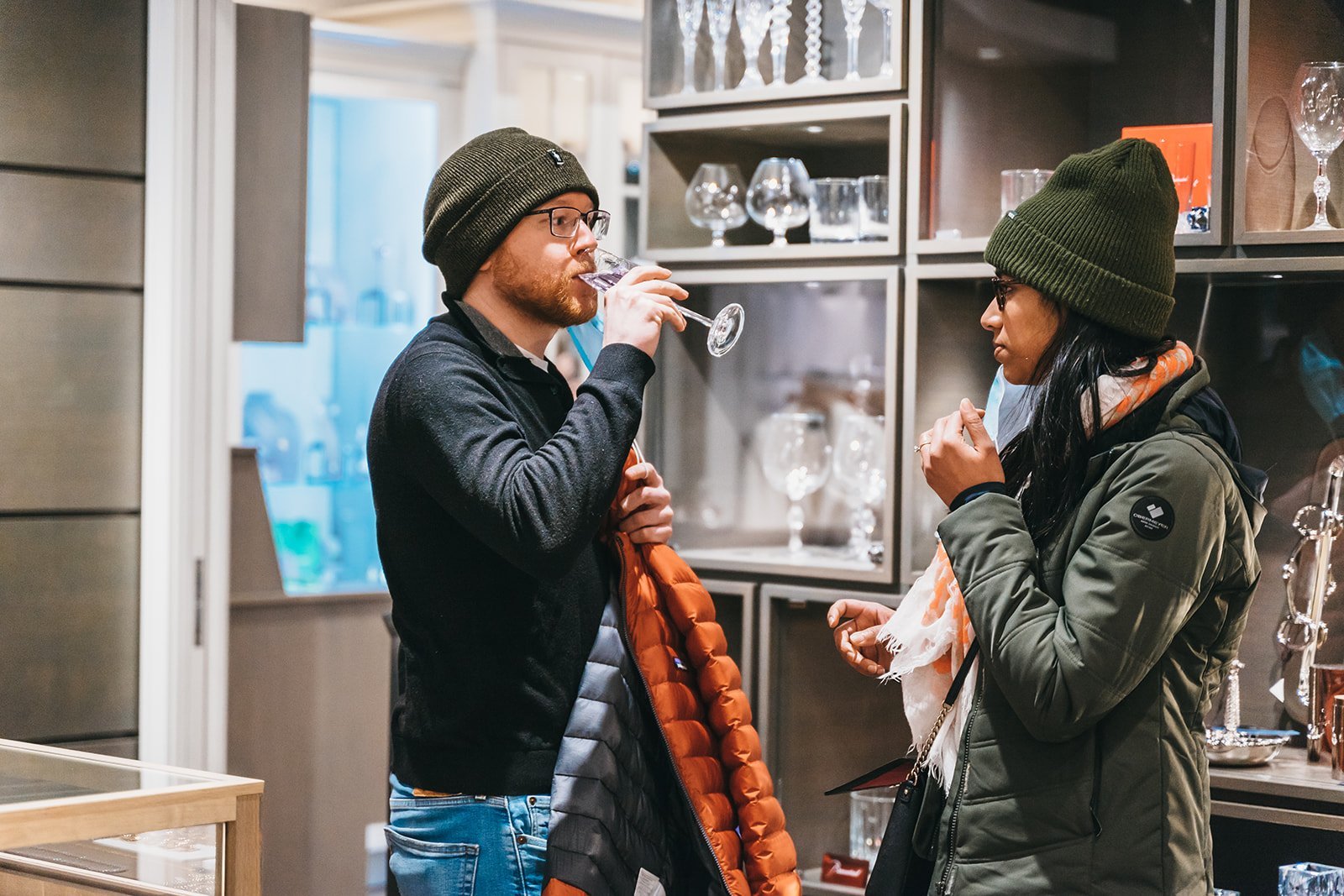 Two people wearing winter clothing, one drinking from a wine glass, standing in front of a display of glassware in a store.