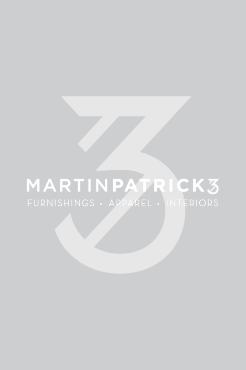 Black and white photo of a smiling person with long hair, wearing a casual blazer over a white blouse and dark jeans, standing against a white brick wall. Greta MartinPatrick 3. Interiors Team.