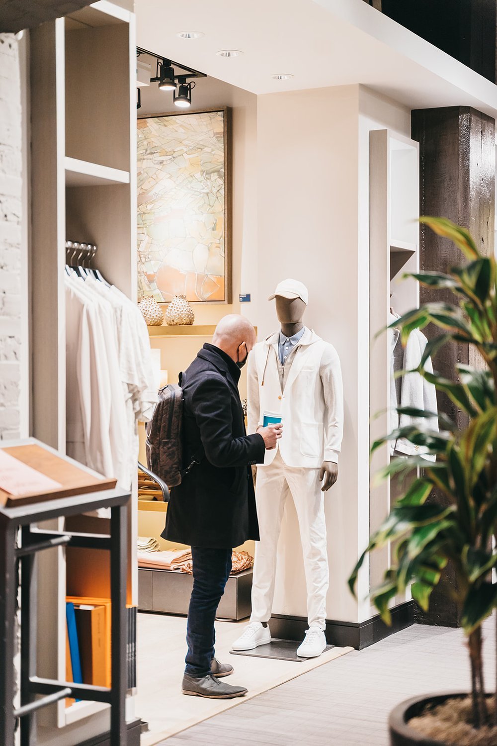 Man in store looking at a mannequin wearing white clothing, next to hanging white shirts and a plant.