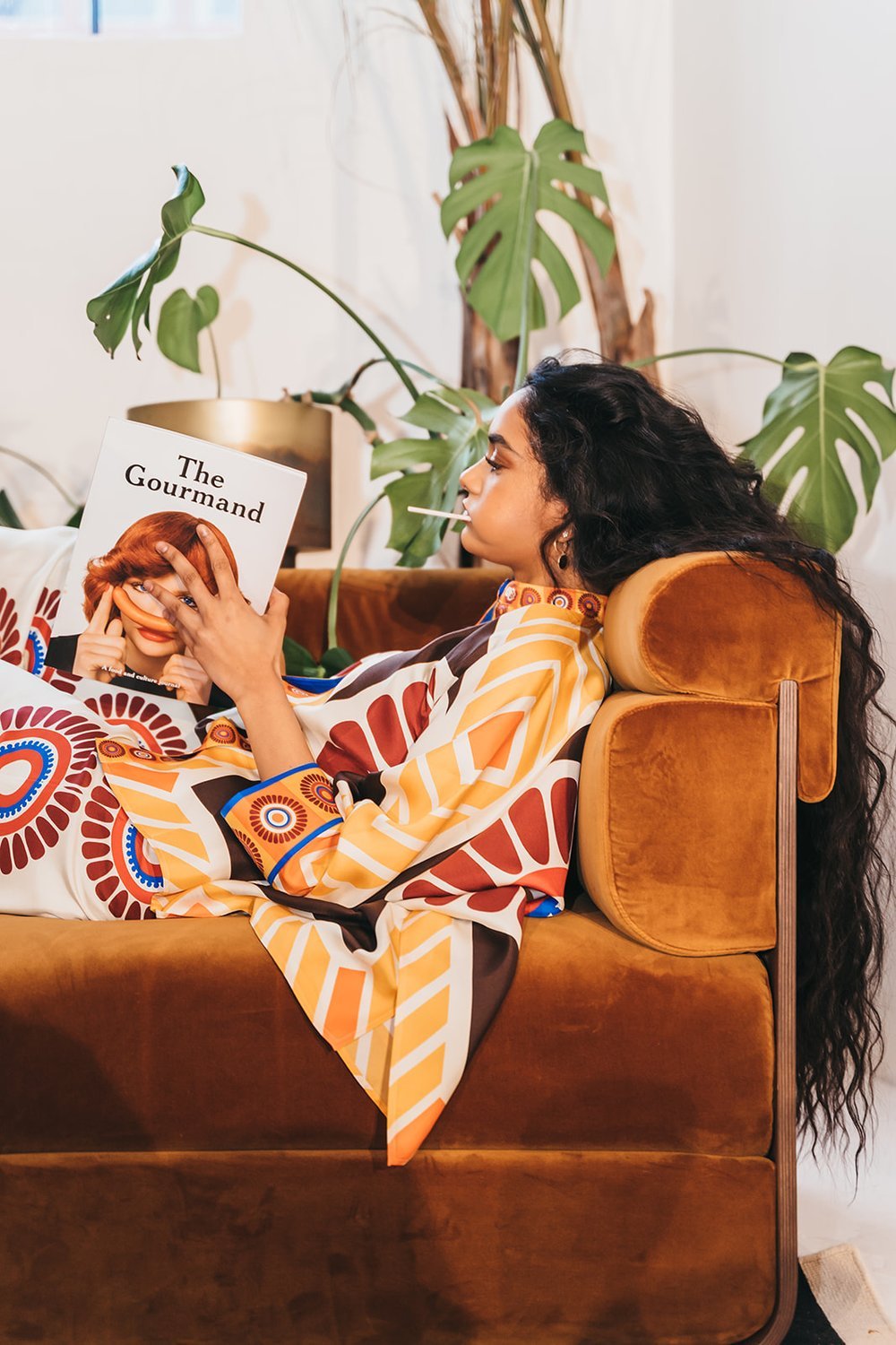 Woman reading a magazine titled 'The Gourmand' while lying on a brown sofa, surrounded by green plants, wearing a colorful patterned outfit with a lollipop in her mouth.