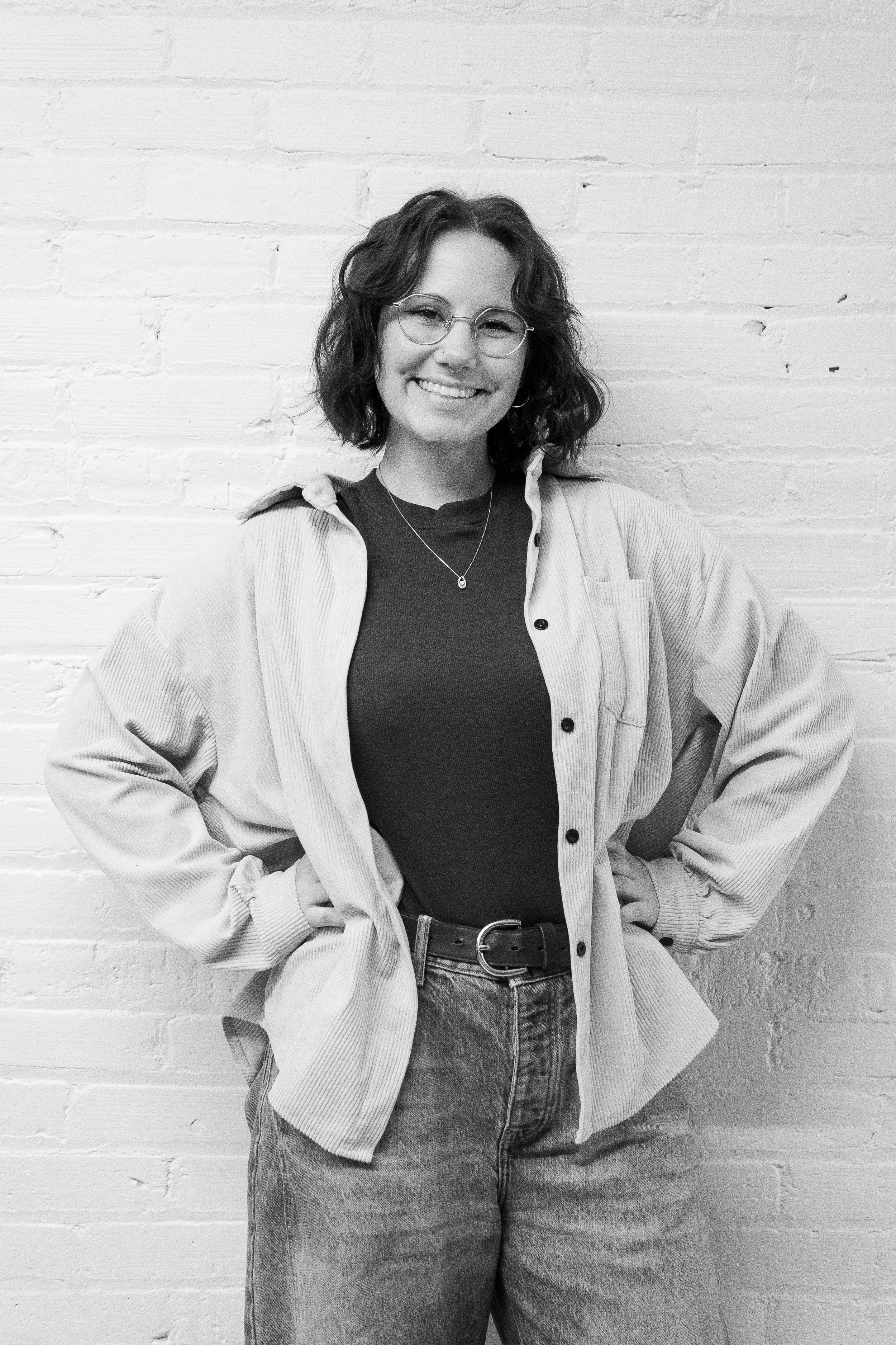 Black and white photo of a smiling person with long hair, wearing a casual blazer over a white blouse and dark jeans, standing against a white brick wall. Greta MartinPatrick 3. Interiors Team.