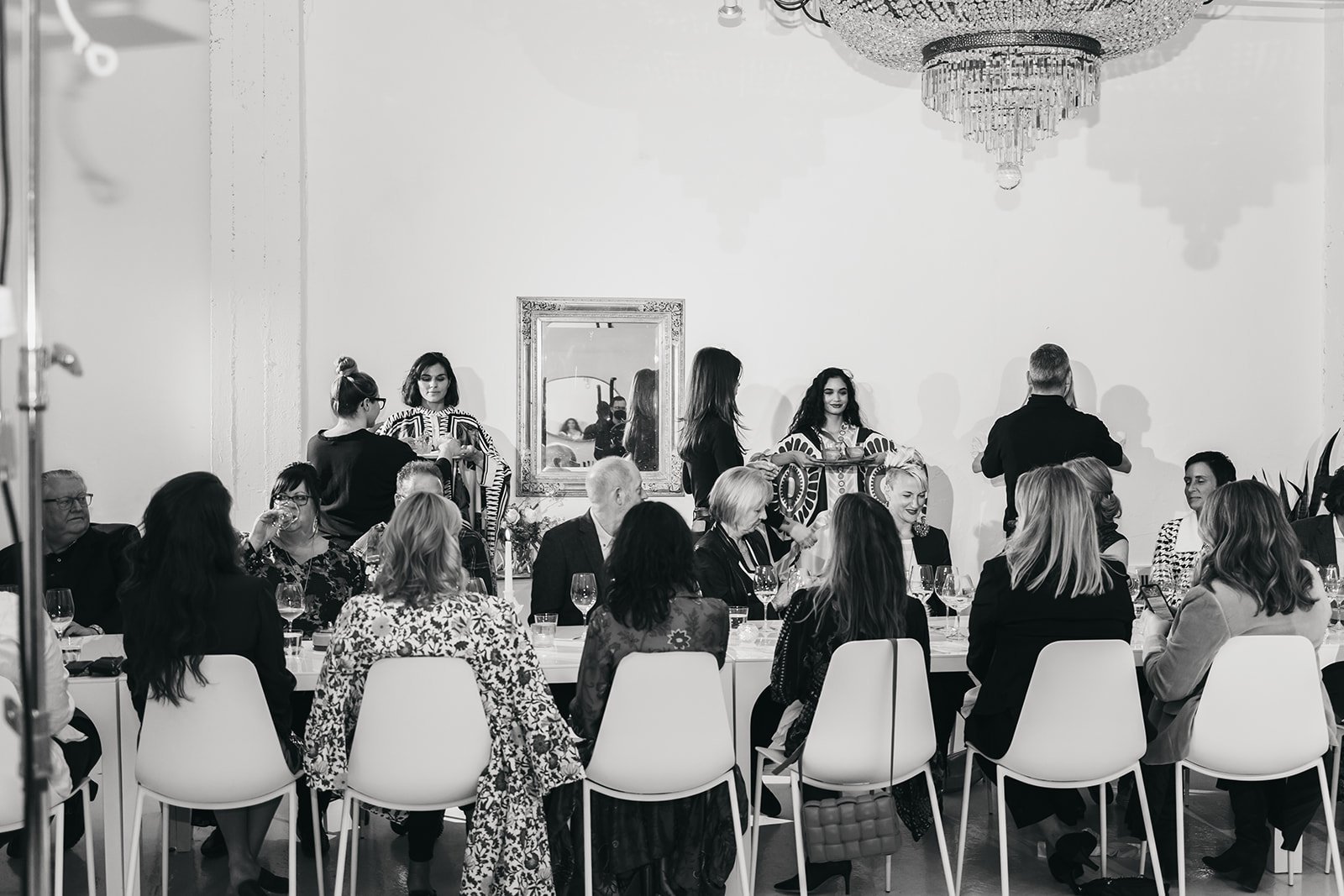Black and white photo of a formal dinner event with seated guests at a long table, servers standing, and a large chandelier overhead.