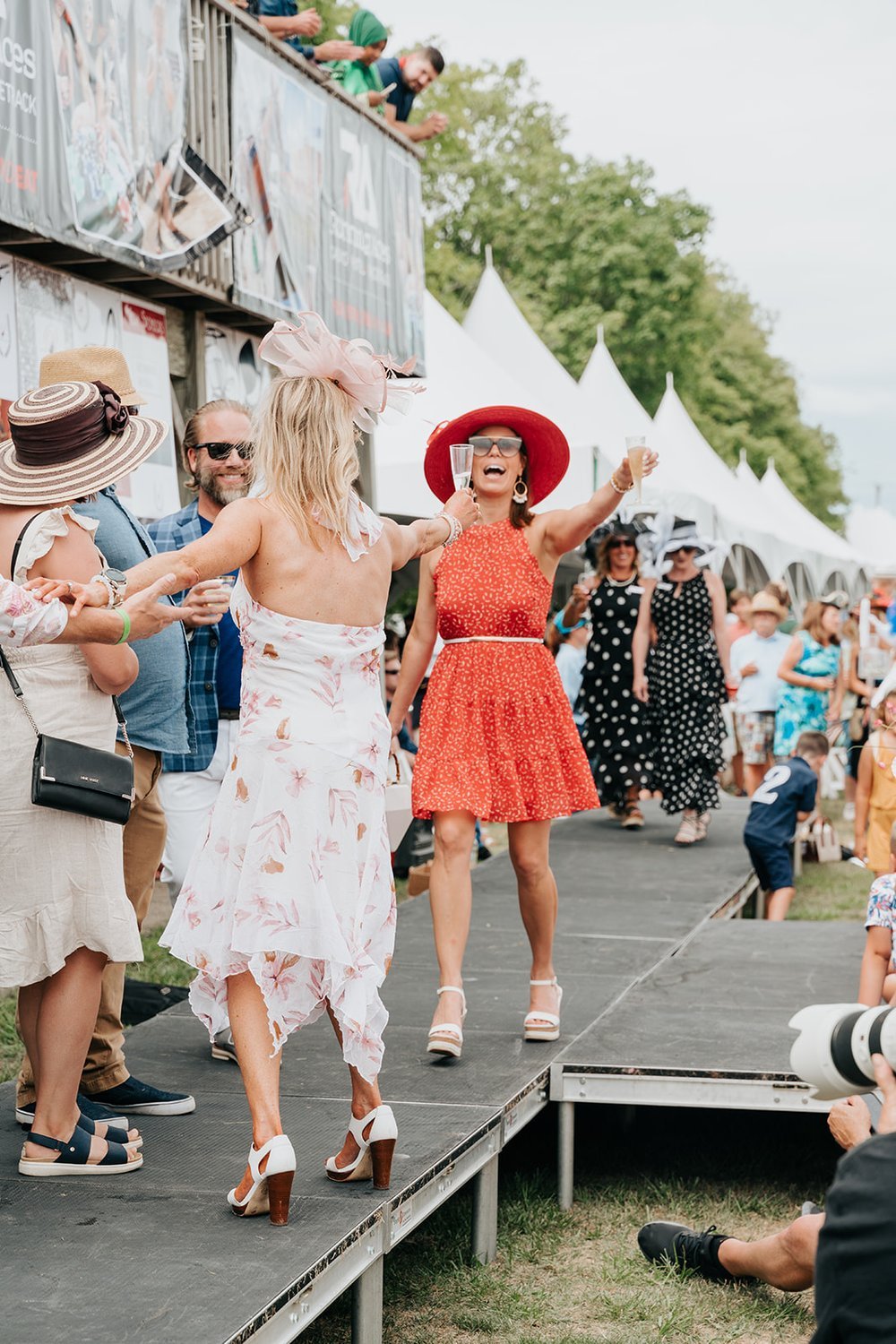 A festive outdoor event with people stylishly dressed, wearing hats. A woman in a red dress and hat raises a glass, smiling. Tents and spectators in the background suggest a lively, social gathering, likely a fashion or horse racing event.