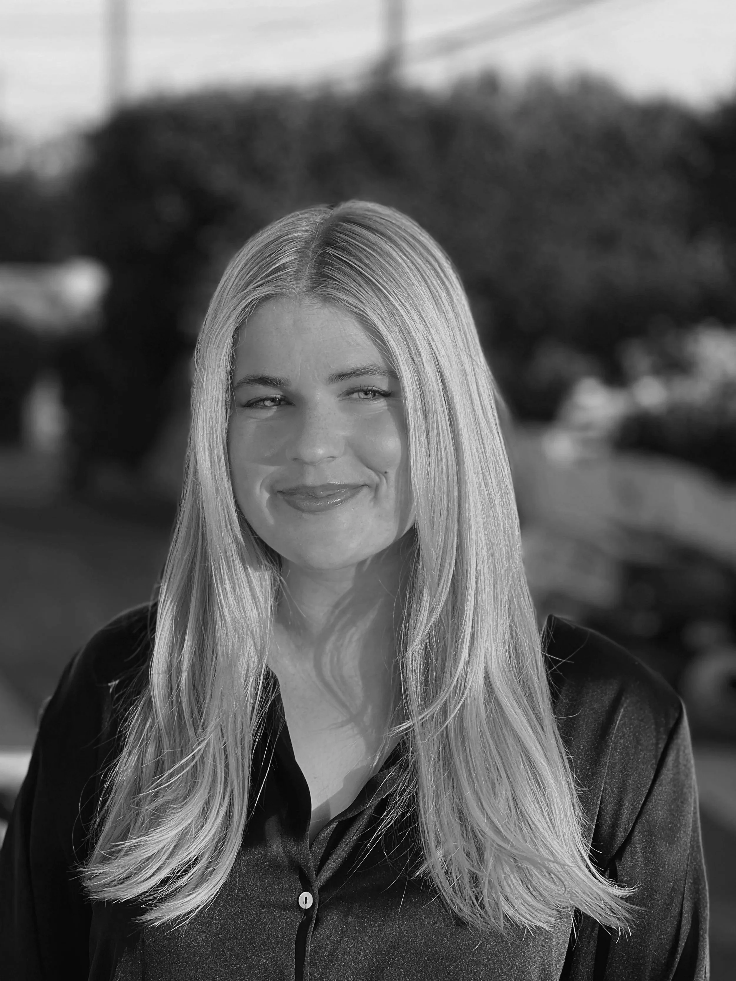 A black and white photo of a woman with long straight hair, smiling slightly, standing outdoors.