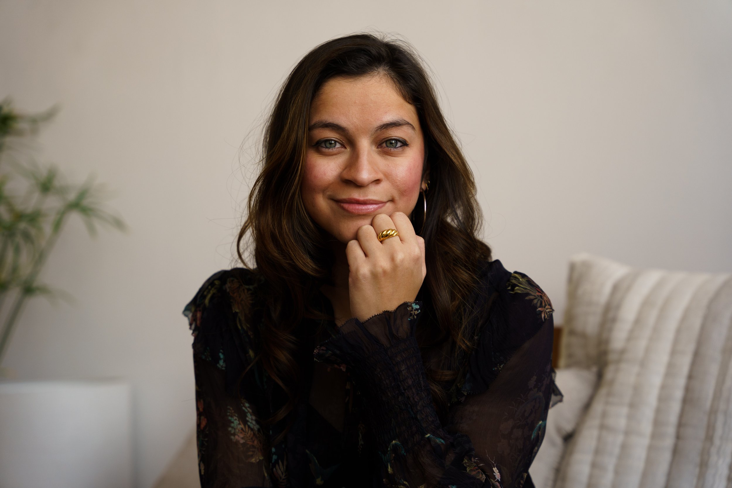A woman with wavy brown hair sits on a beige couch, wearing a black floral top, smiling with her head tilted slightly and resting her chin on her hand that has a gold ring.