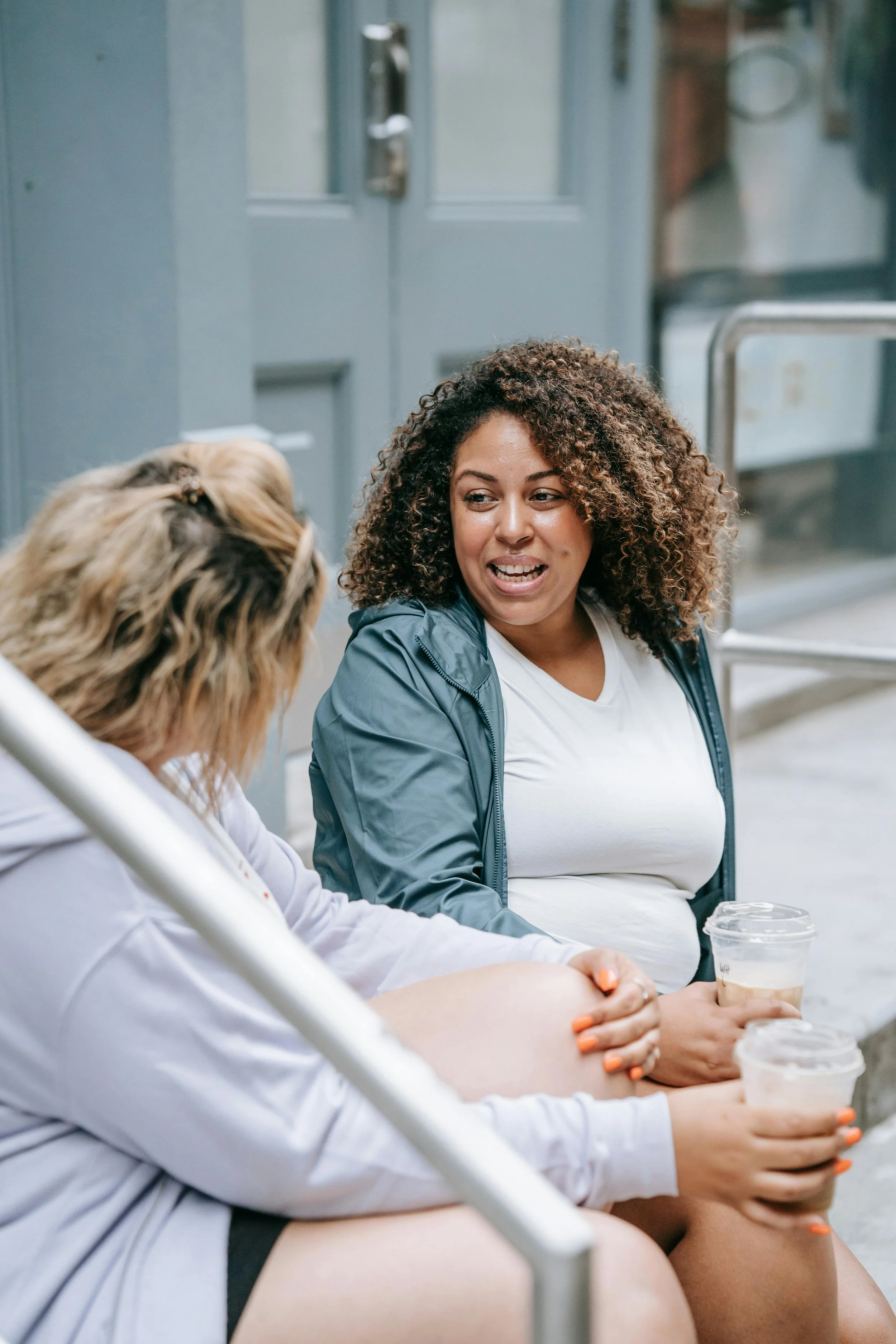 Two women sitting outside, having a conversation. One woman with curly hair, wearing a gray jacket, smiling and holding a coffee cup. The other woman with light-colored hair, wearing a white hoodie, resting her hand on her knee.