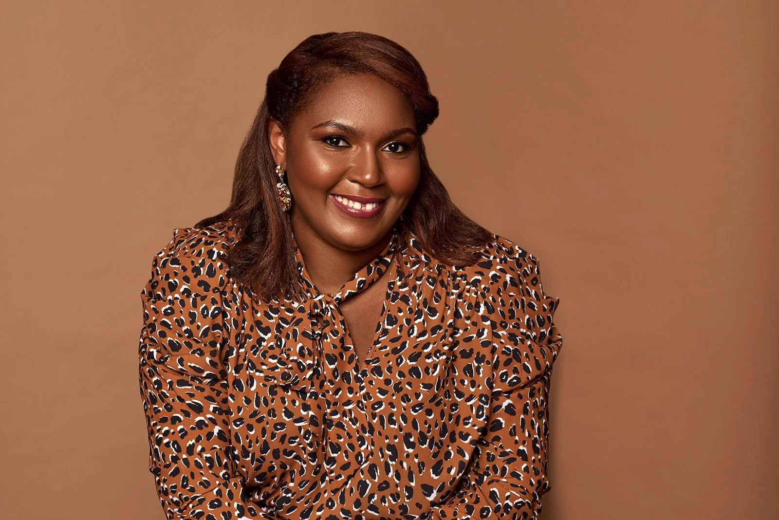 Portrait of a smiling woman with brown hair, wearing a leopard print blouse and earrings, against a brown background.