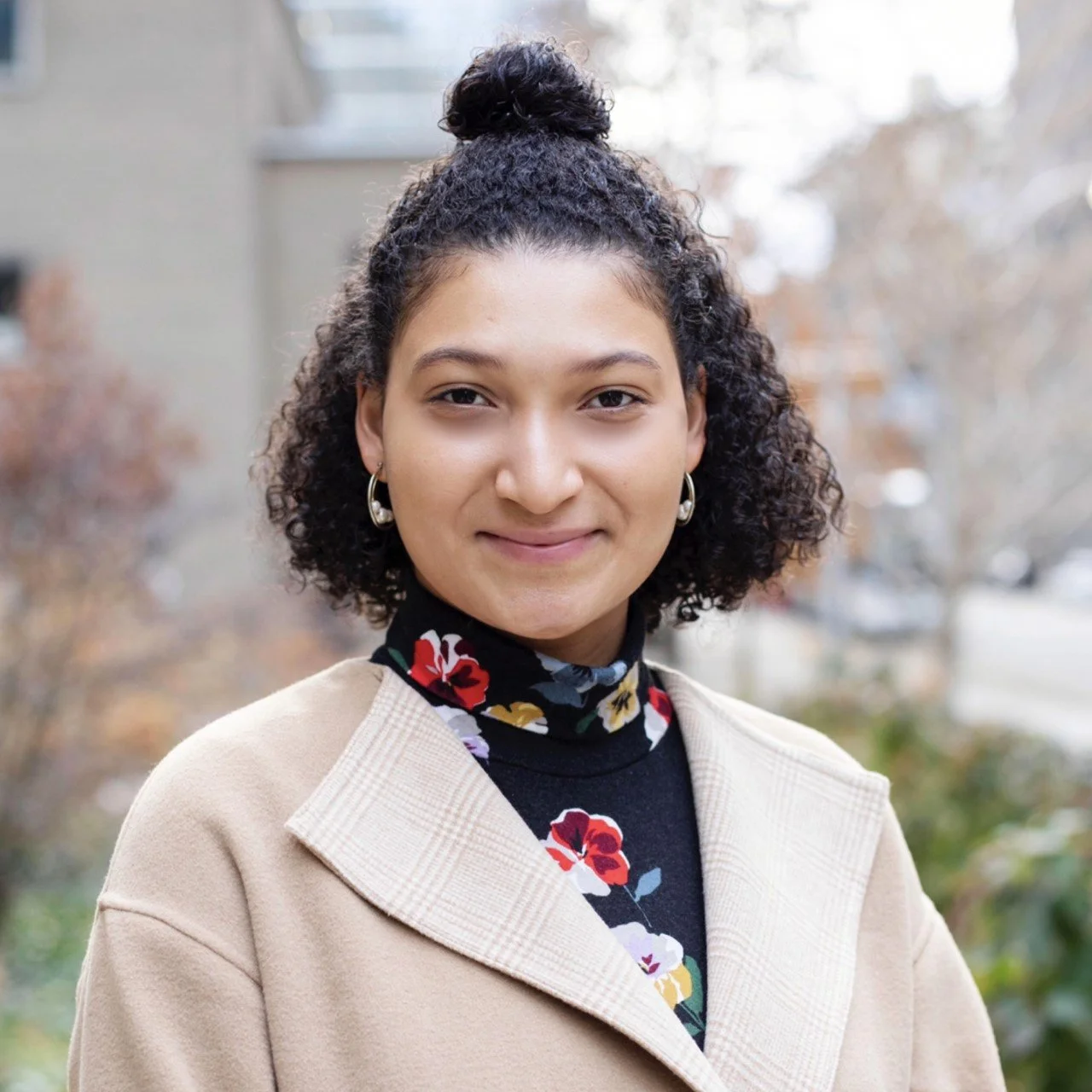 A young woman with curly hair in a bun, wearing hoop earrings, a floral turtleneck, and a beige coat, standing outdoors with trees and a building in the background.