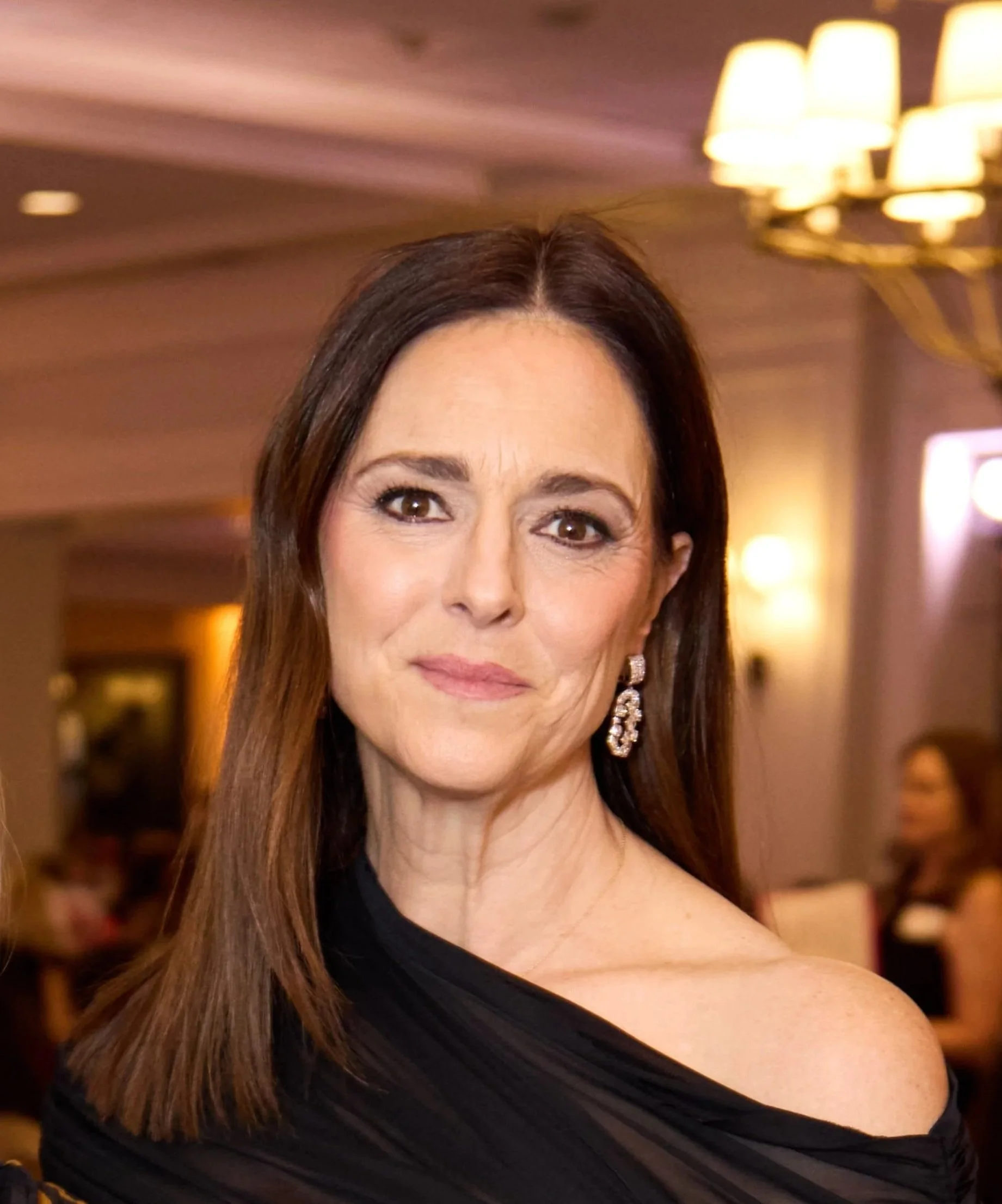 Close-up portrait of a woman with brown hair, wearing earrings, looking at the camera with a slight smile.