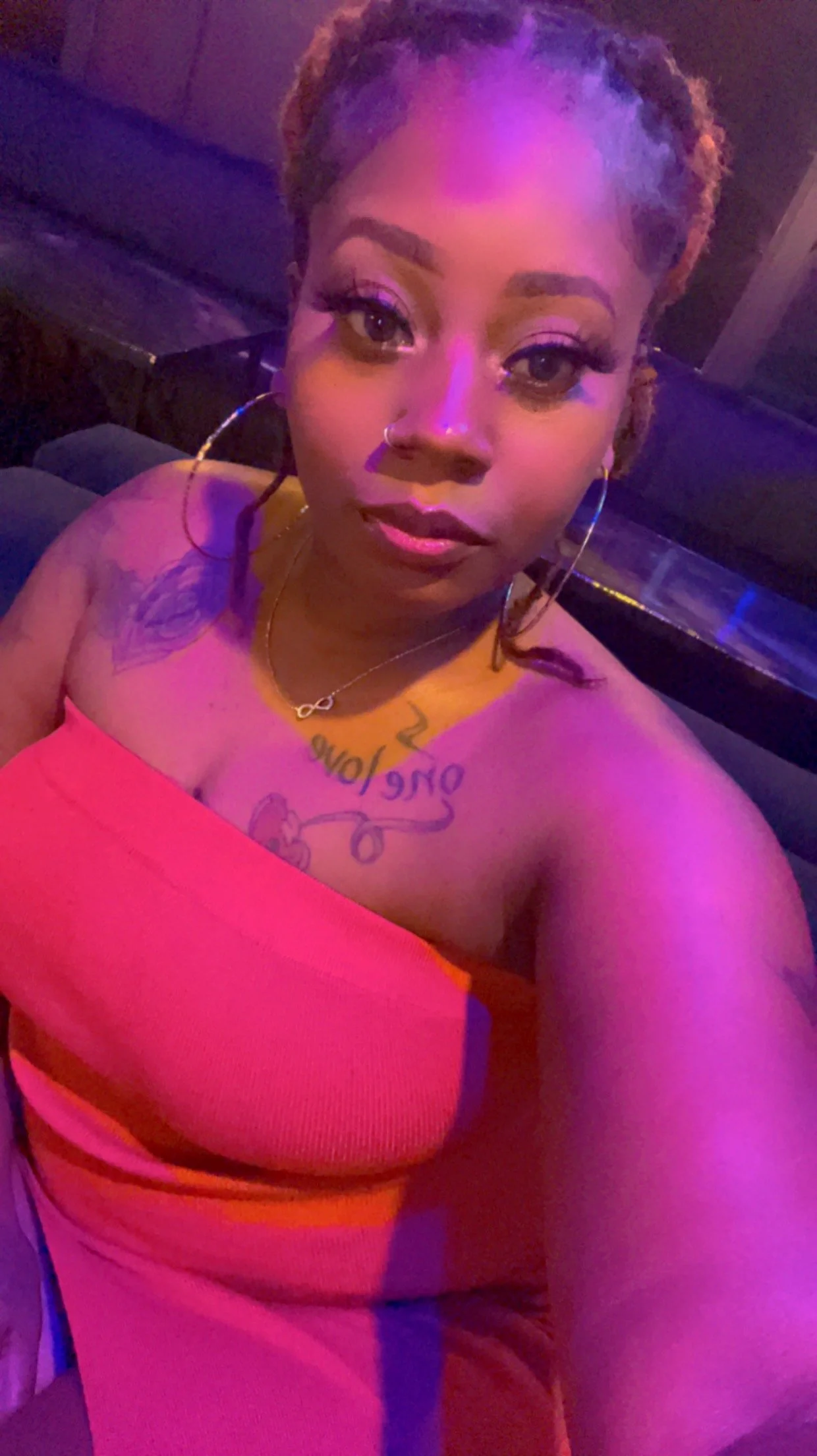 A woman with tattoos and hoop earrings taking a selfie in a dimly lit setting with purple lighting. She is wearing a strapless pink top and a gold necklace.