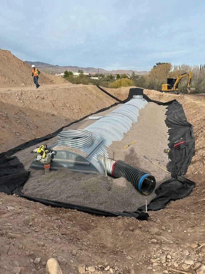 Construction site with large metal pipe and black liner, worker in safety gear, and small yellow excavator in background.