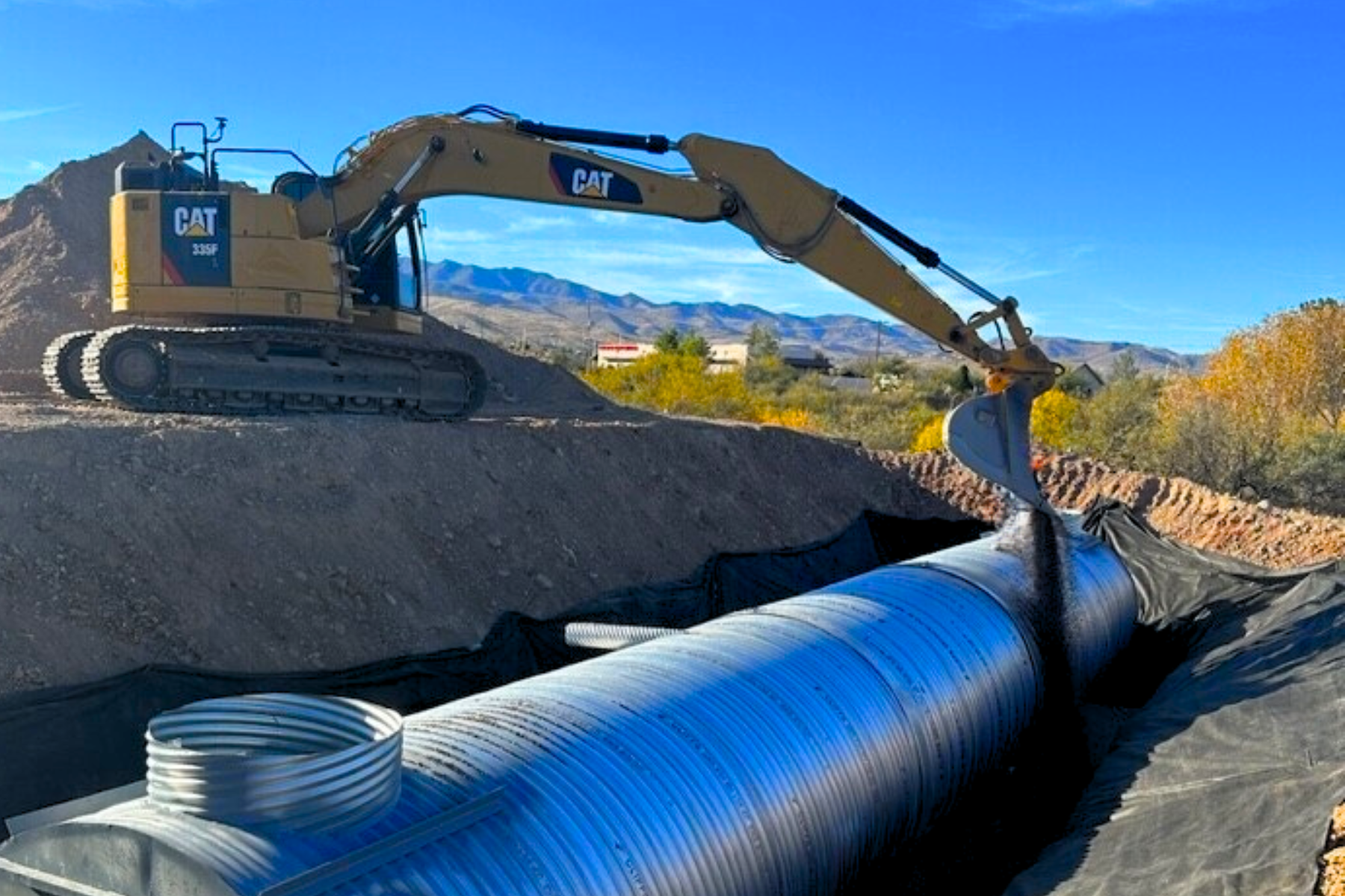A construction excavator with a Caterpillar logo is digging a trench for underground pipes in an outdoor setting with mountains and trees in the background.