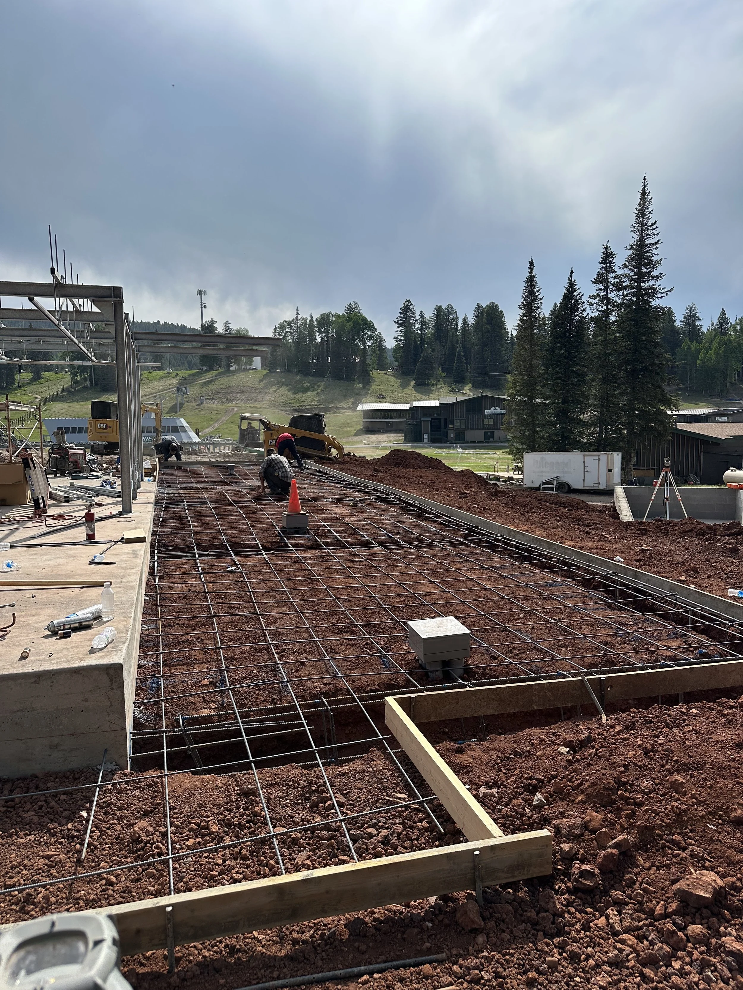 Construction workers laying rebar on a building foundation with mountains and trees in the background.