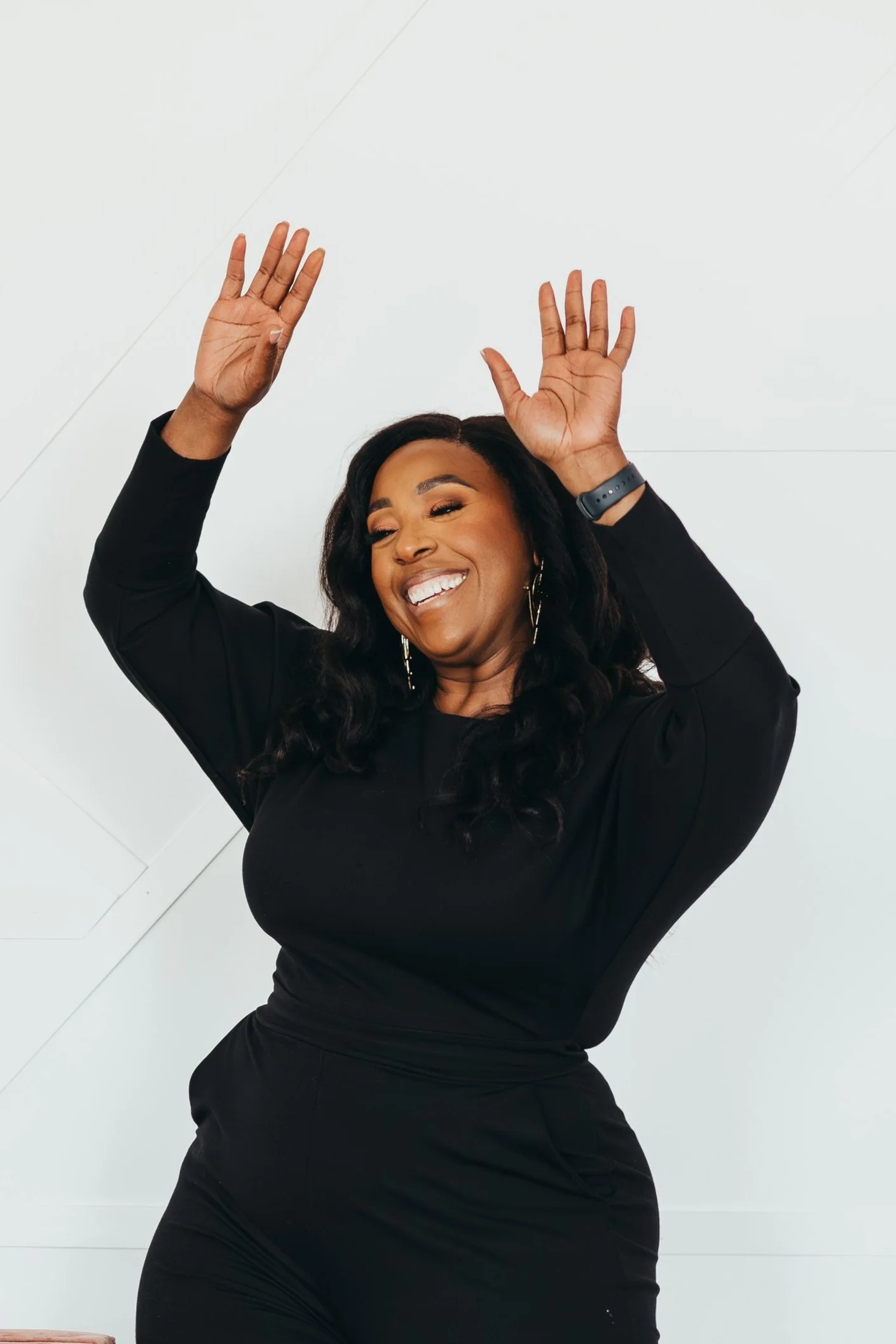 A woman with long dark hair, wearing a black long-sleeve dress and earrings, is smiling and dancing with her hands raised in the air.