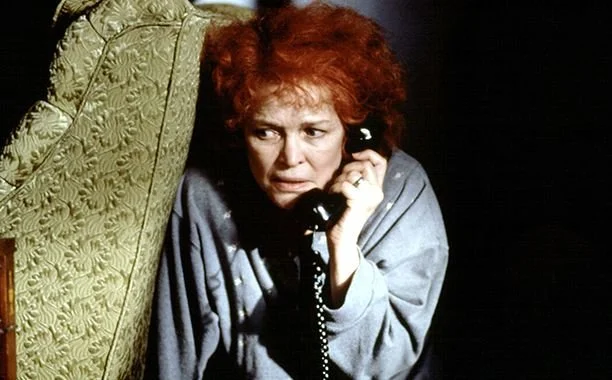 A woman with red curly hair holding a black landline phone to her ear, looking worried, sitting beside a green, patterned armchair.