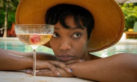 Person wearing a wide-brimmed hat resting their chin on a poolside edge next to a cocktail glass with a cherry inside.
