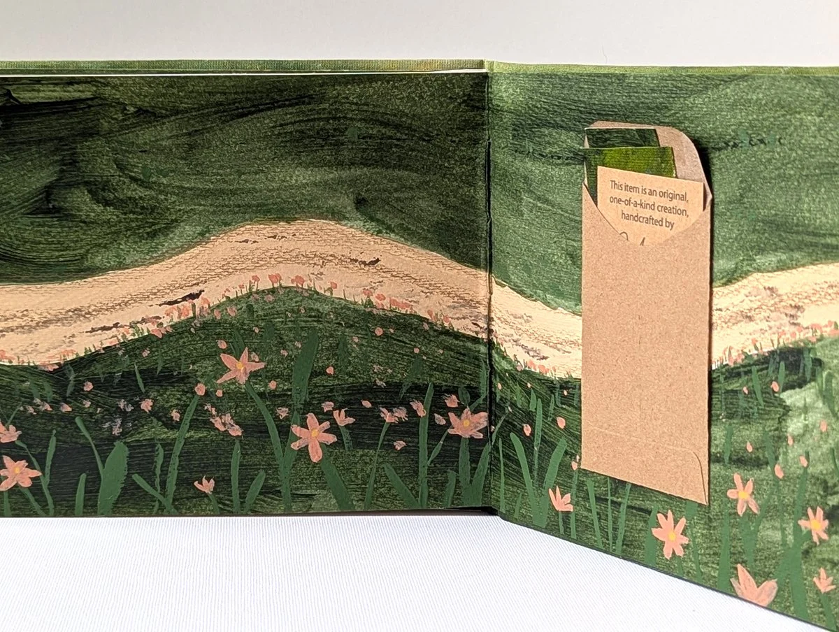 Open handcrafted pop-up card with a painted landscape of green hills, pink flowers, a winding path, and a box-shaped pocket on the right side containing a message.