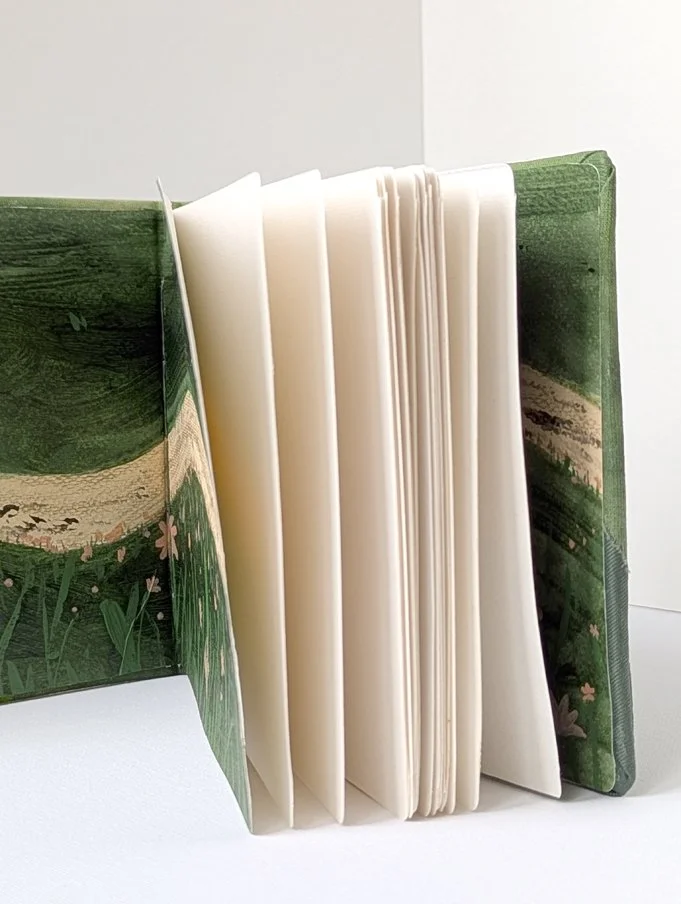 Open children's book with white pages, standing upright on a white surface, with a green cover featuring a nature scene.