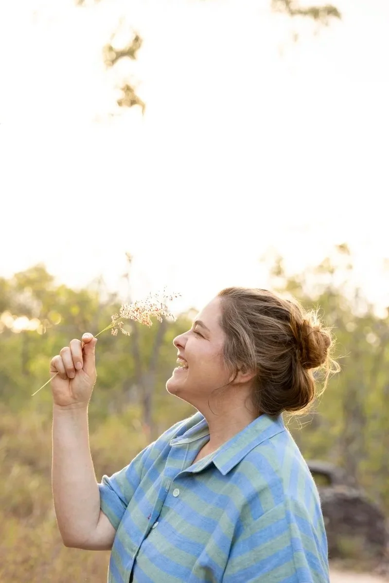 A woman (Brenner Lowe) smiling and holding a sprig of grass in a natural outdoor setting with trees in the background.