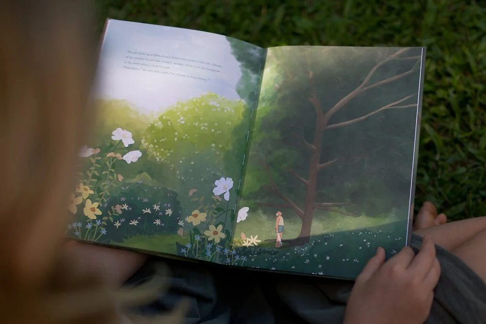 A hand holding a picturebook, showing a double-page drawing of a girl looking up into a tall tree. There are colourful flowers in the garden that surrounds the girl.