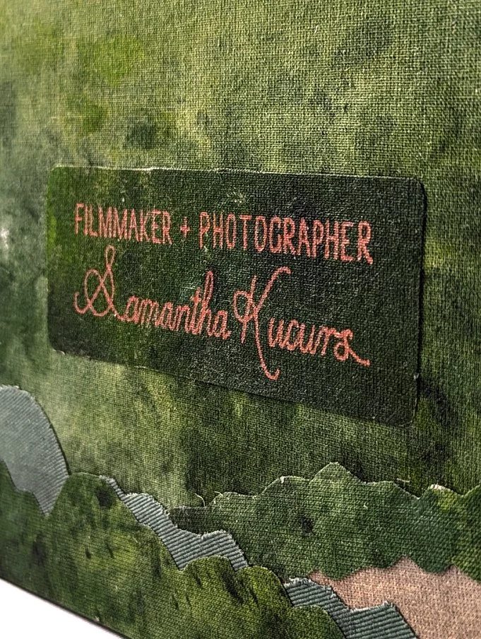 Close-up image of a fabric label on a green and leafy textured background, reading 'Filmmaker + Photographer Samantha Kucurs'.