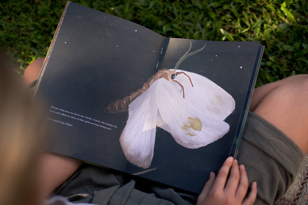 A woman sits on grass with a picture storybook open on a page with a large moth drawn on a dark night sky background.