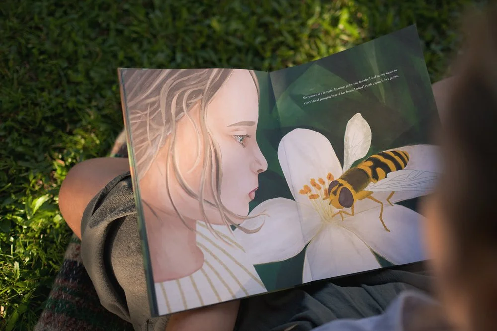 Woman reading a picture storybook with a drawing of a girl's face as she looks at a hoverfly on a white flower.
