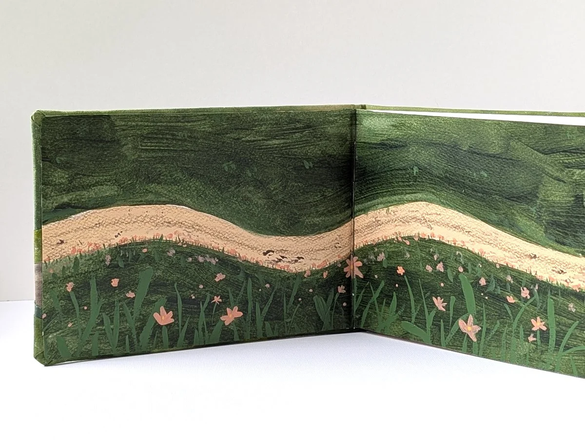 Open illustrated children's book showing a grassy landscape with pink flowers, a sandy path, and rolling green hills.