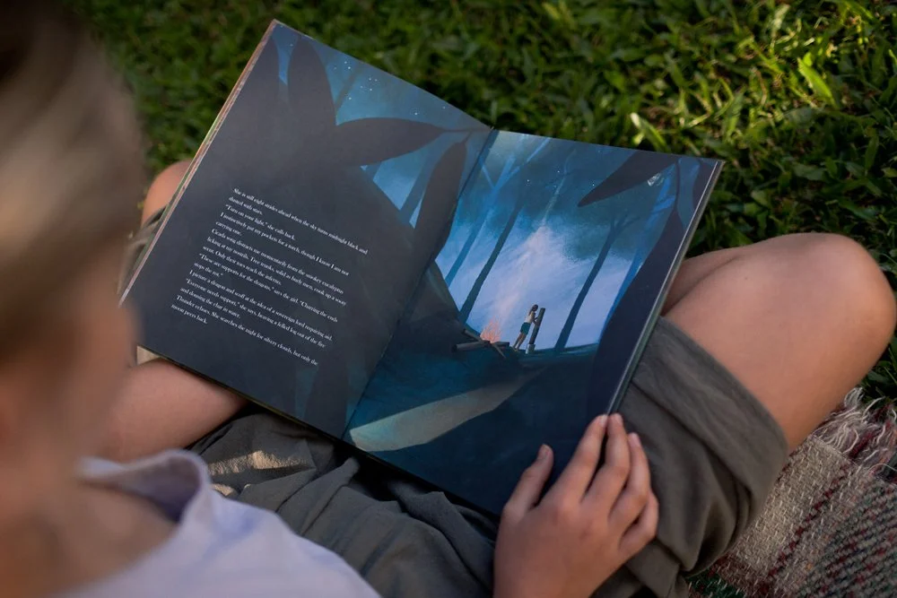 A woman reading a picture book. The book is open to a dark forest and a girl standing besides a campfire. Smoke rises upwards.
