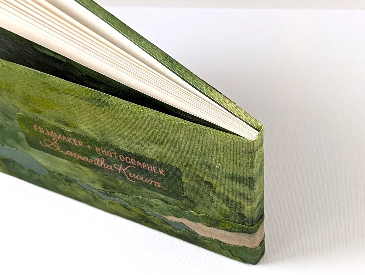 Close-up of a green book with 'Filmmaker & Photographer, Samantha Kucurs' written on the back cover, with a landscape scene of green hills.