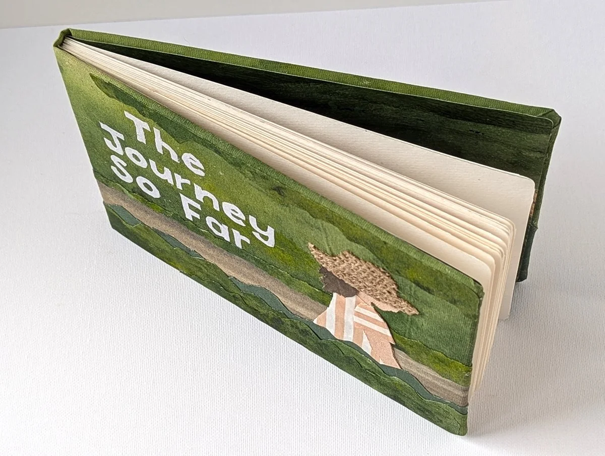 A colorful book with the title "The Journey So Far" displayed on the cover, featuring an illustrated landscape scene with trees and a pathway.