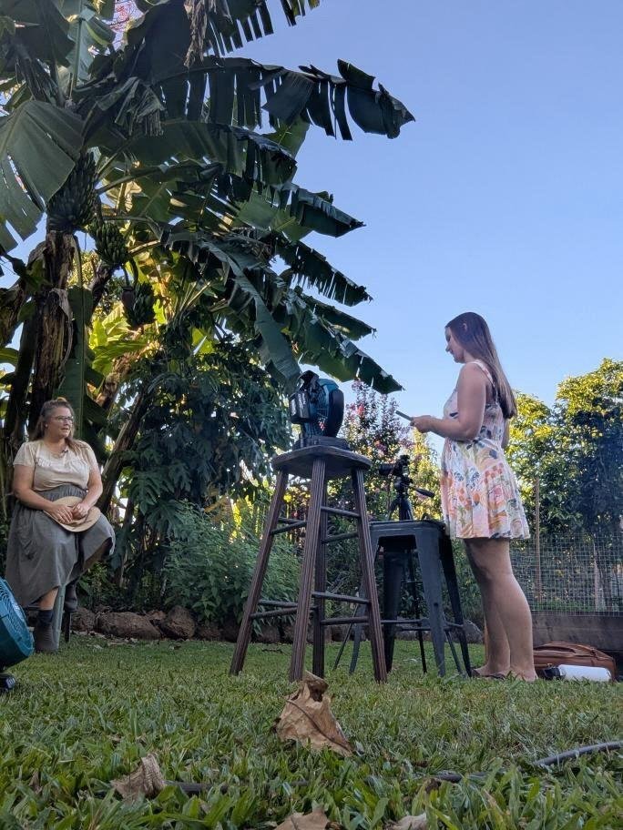 Two women outdoors in a lush garden with large banana leaves and trees. One woman, seated on a chair, wears a beige top and gray skirt. The other woman, standing, wears a floral dress and is looking at her phone. There is camera equipment set up, including a camera on a tripod, and a tall stool with additional equipment on it.