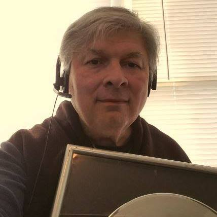A man with gray hair wearing a headset and a dark shirt, holding a computer monitor, in a room with beige walls and closed blinds.