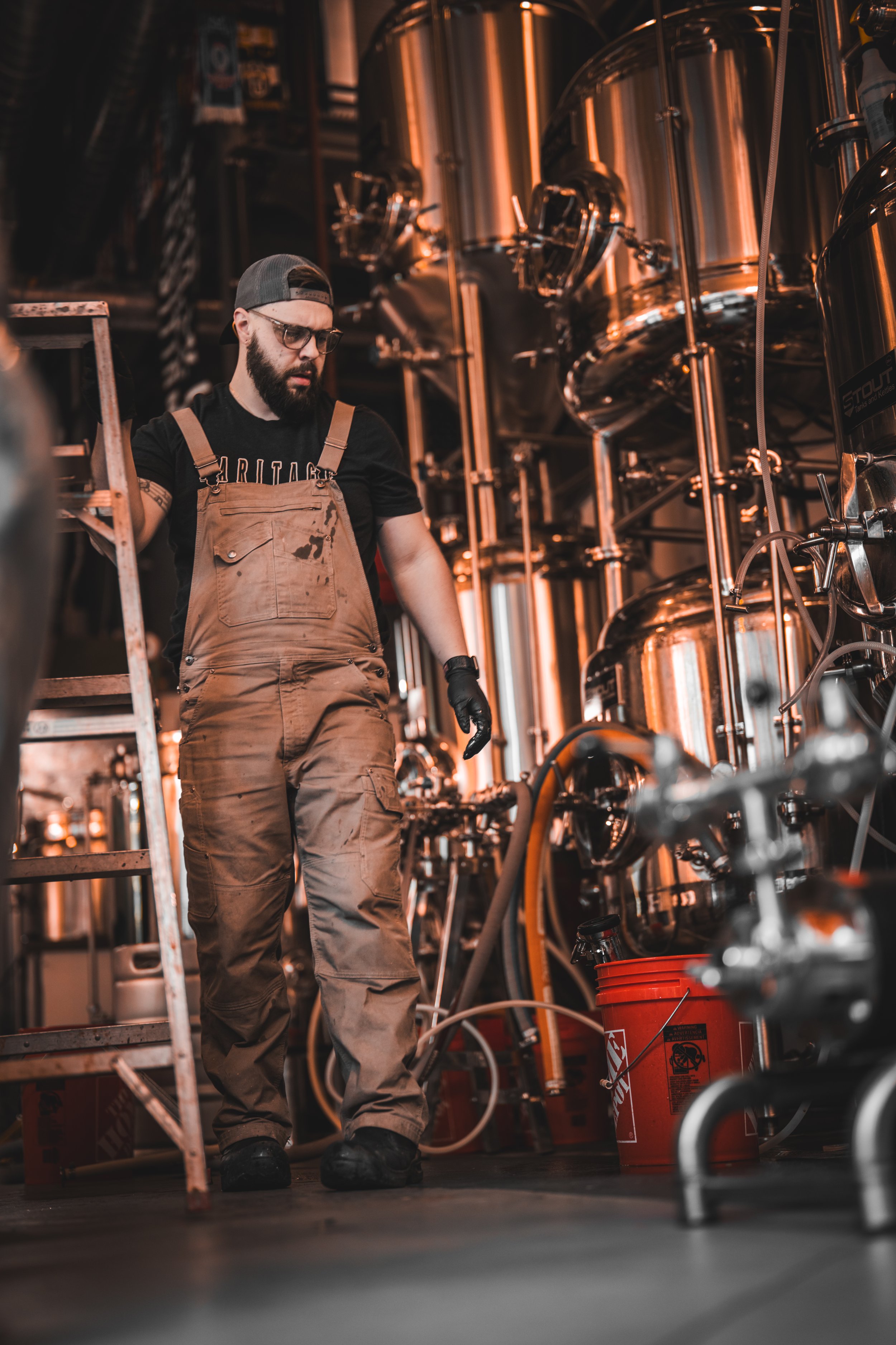 A man in protective gloves and glasses wearing work overalls manages brewing equipment in a brewery.
