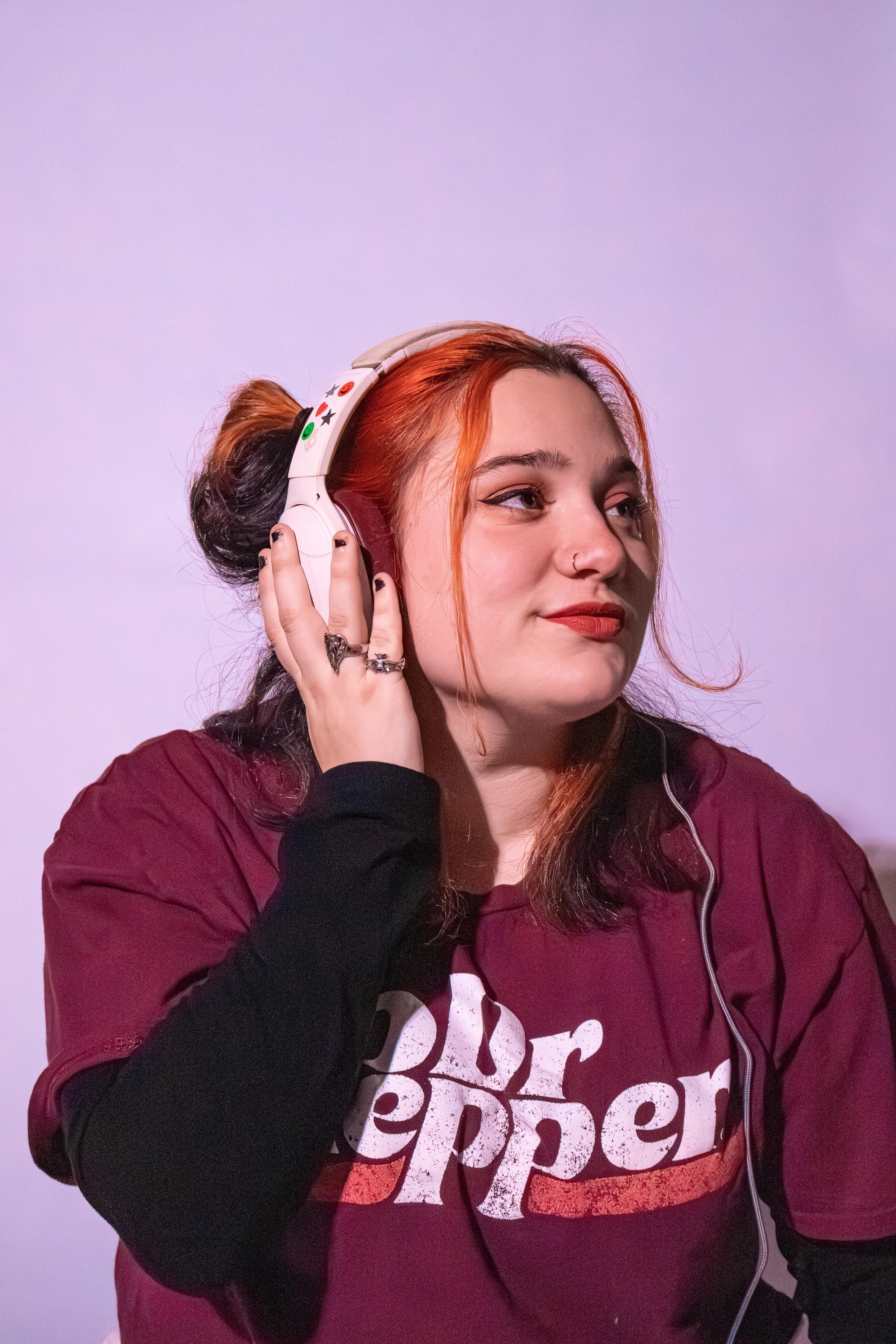 A young woman with red hair in a bun, wearing headphones and a maroon 'Dr Pepper' t-shirt, listening to music.