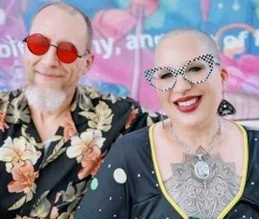 Two people smiling at a colorful event, one wearing red sunglasses and a floral shirt, the other with patterned glasses and visible tattoos.