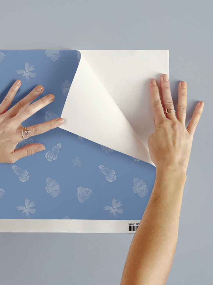 Hands applying blue peel-and-stick wallpaper featuring a soft watercolour seashell pattern in a coastal style. Close-up application mockup showing the design and texture against a light wall