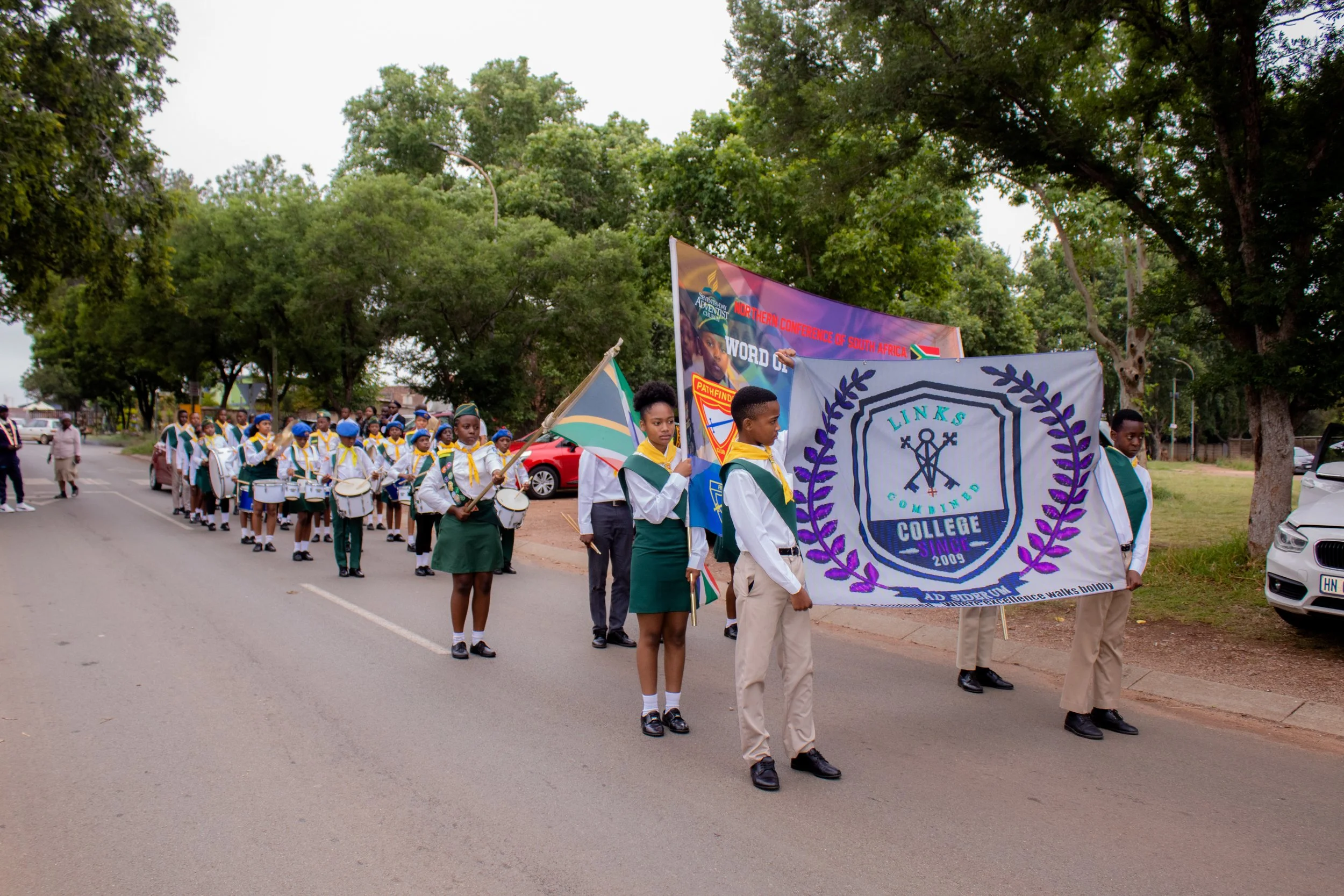 A parade of school children walking along a street, holding flags and banners, with trees and parked cars on the side.
