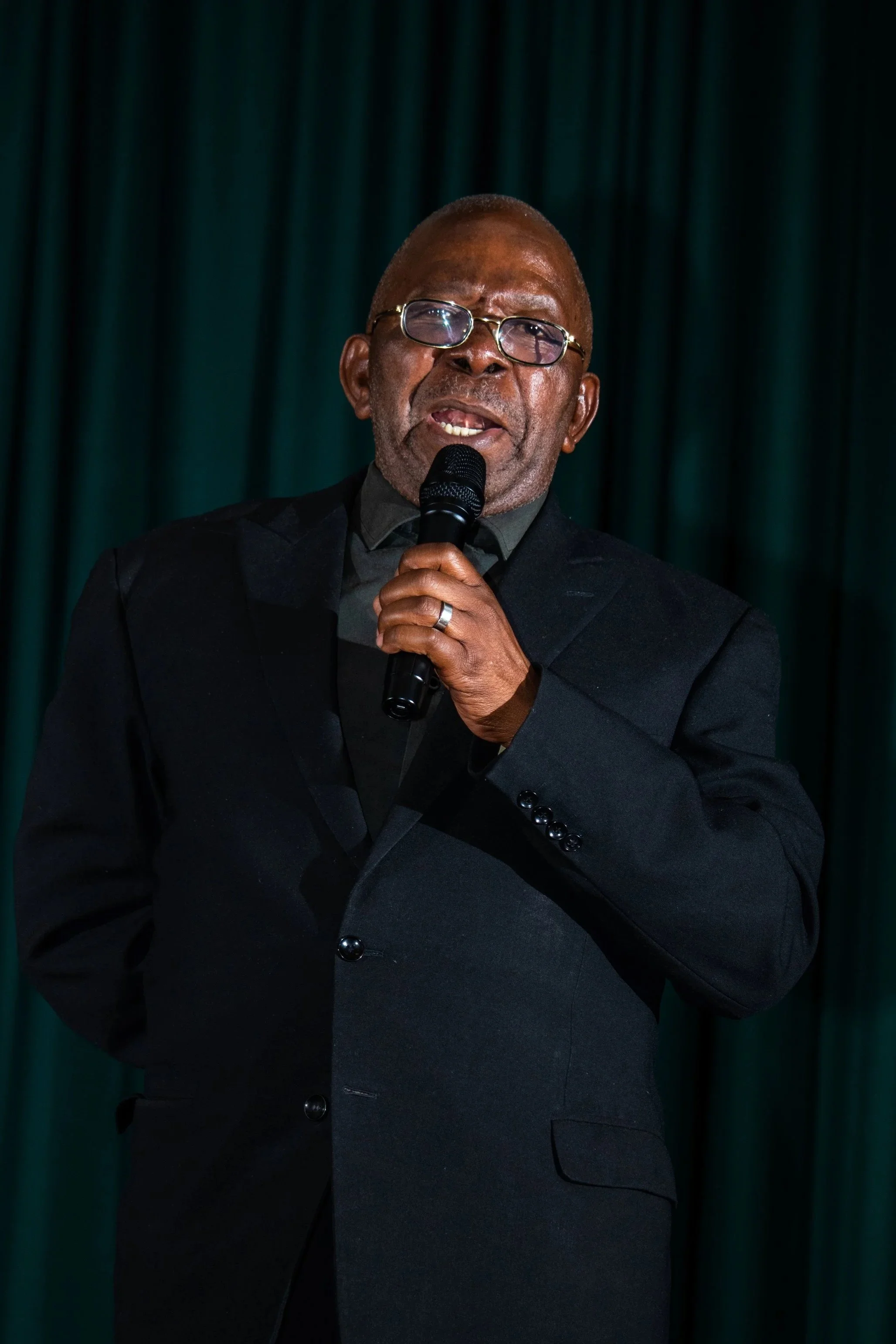 An elderly man with glasses holding a microphone, wearing a dark suit, speaking on stage with a dark green curtain backdrop.