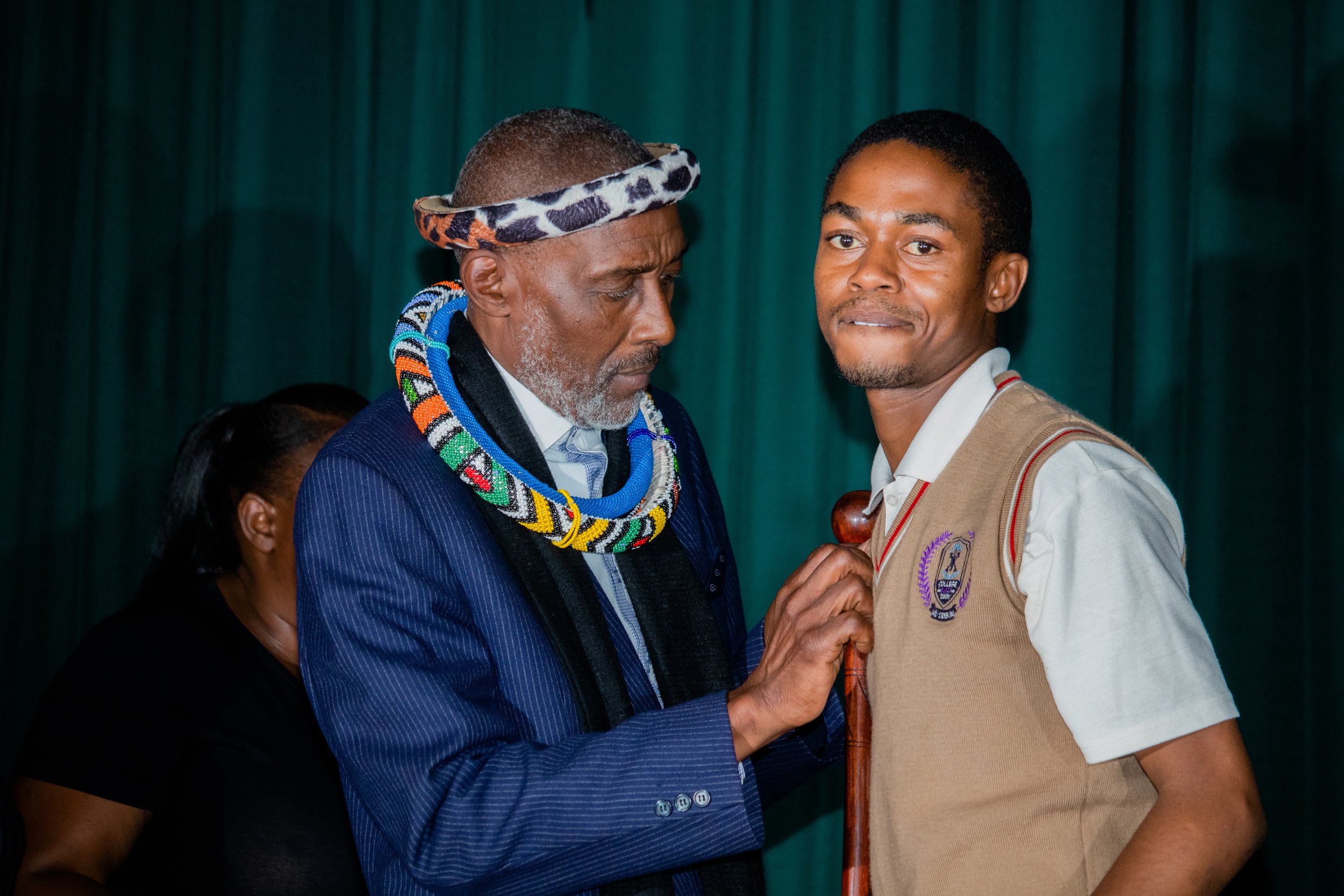 A local South African chief dressed in traditional attire with beads and a headband interacting with a young man in a school uniform.