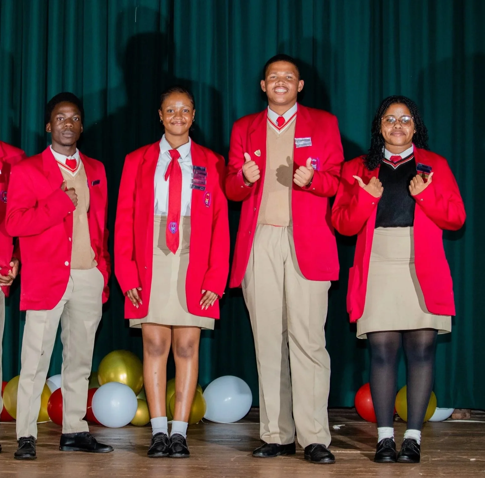 Group of students in red blazers and beige skirts or pants standing on a stage with green curtains and balloons.