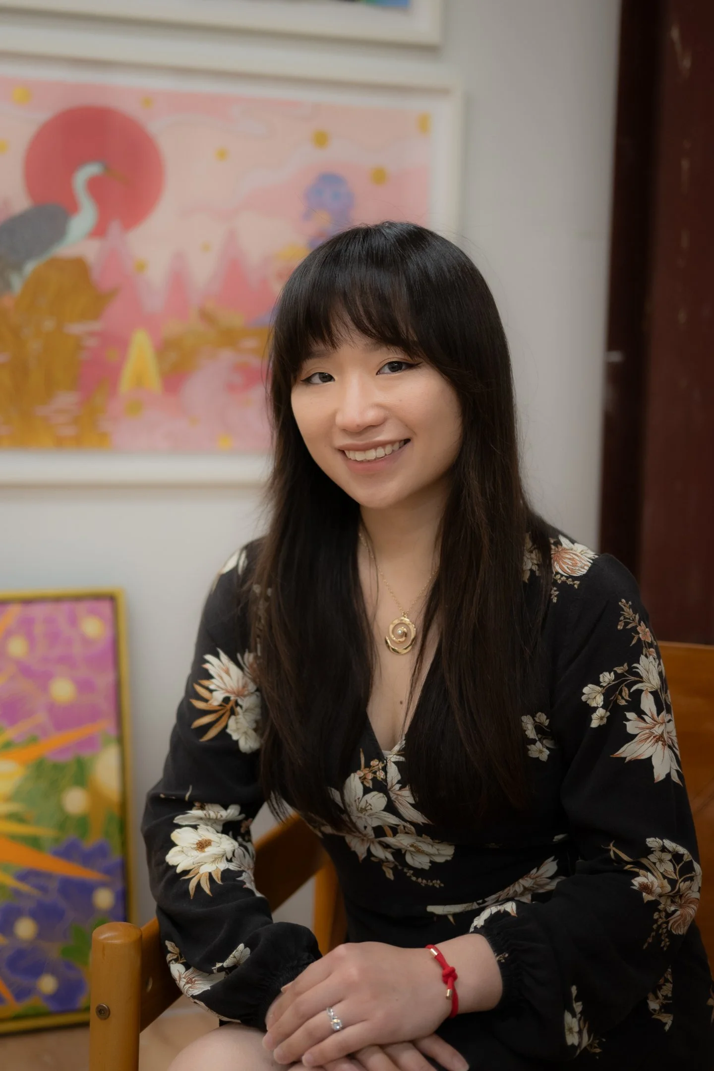 Julie Xiao Headshot. A woman sitting at a table smiling, wearing a black floral dress, gold necklace, and red bracelet, with colorful artwork in the background.