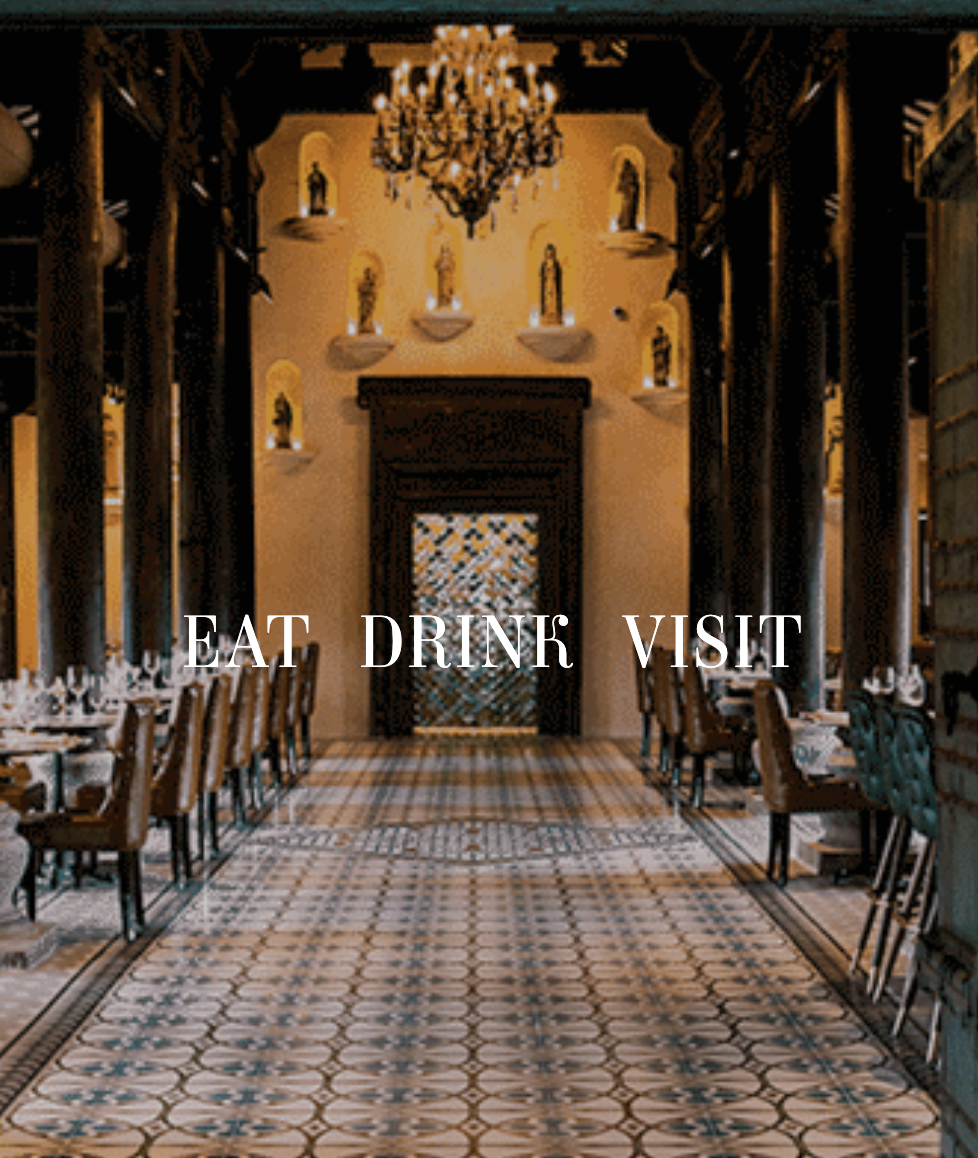 Elegant restaurant interior with a chandelier, wall niches with statues, and a tiled floor. Overlaid text reads 'EAT DRINK VISIT'.