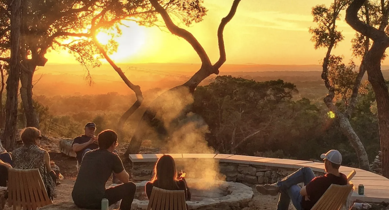 Group of people sitting around a fire pit outdoors at sunset surrounded by trees.